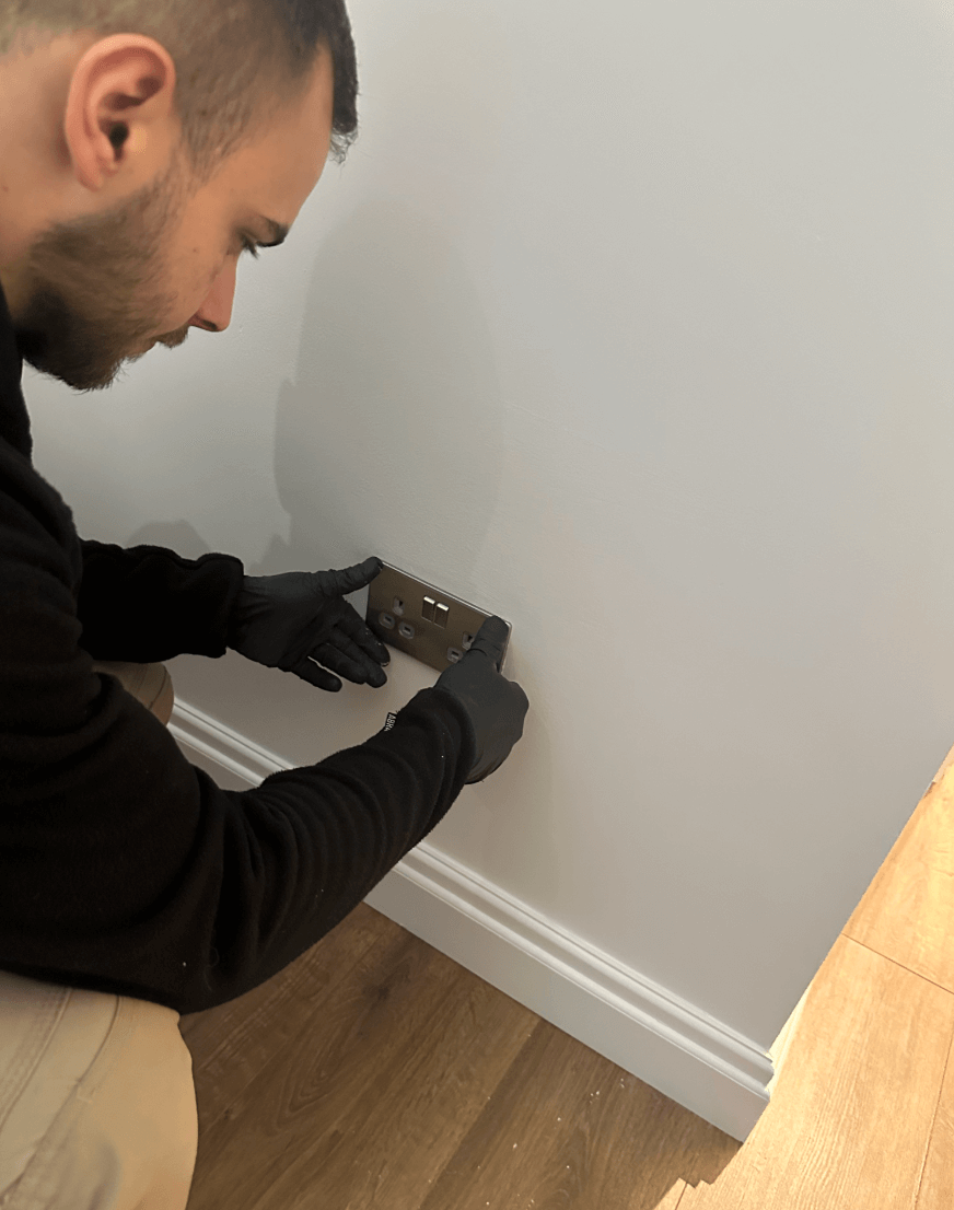 Electrician wearing black gloves installing a brushed steel double socket on a freshly painted white wall in a modern interior with wooden flooring.